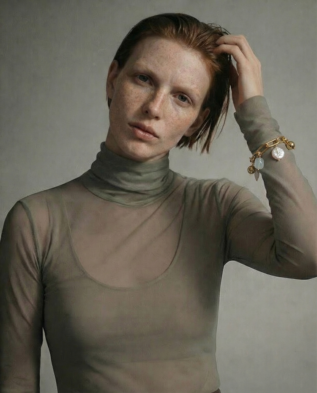 Woman wearing a sheer green turtleneck against a plain background with a gold link charm bracelet on her arm