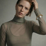Woman wearing a sheer green turtleneck against a plain background with a gold link charm bracelet on her arm