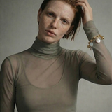 Woman wearing a sheer green turtleneck against a plain background with a gold link charm bracelet on her arm