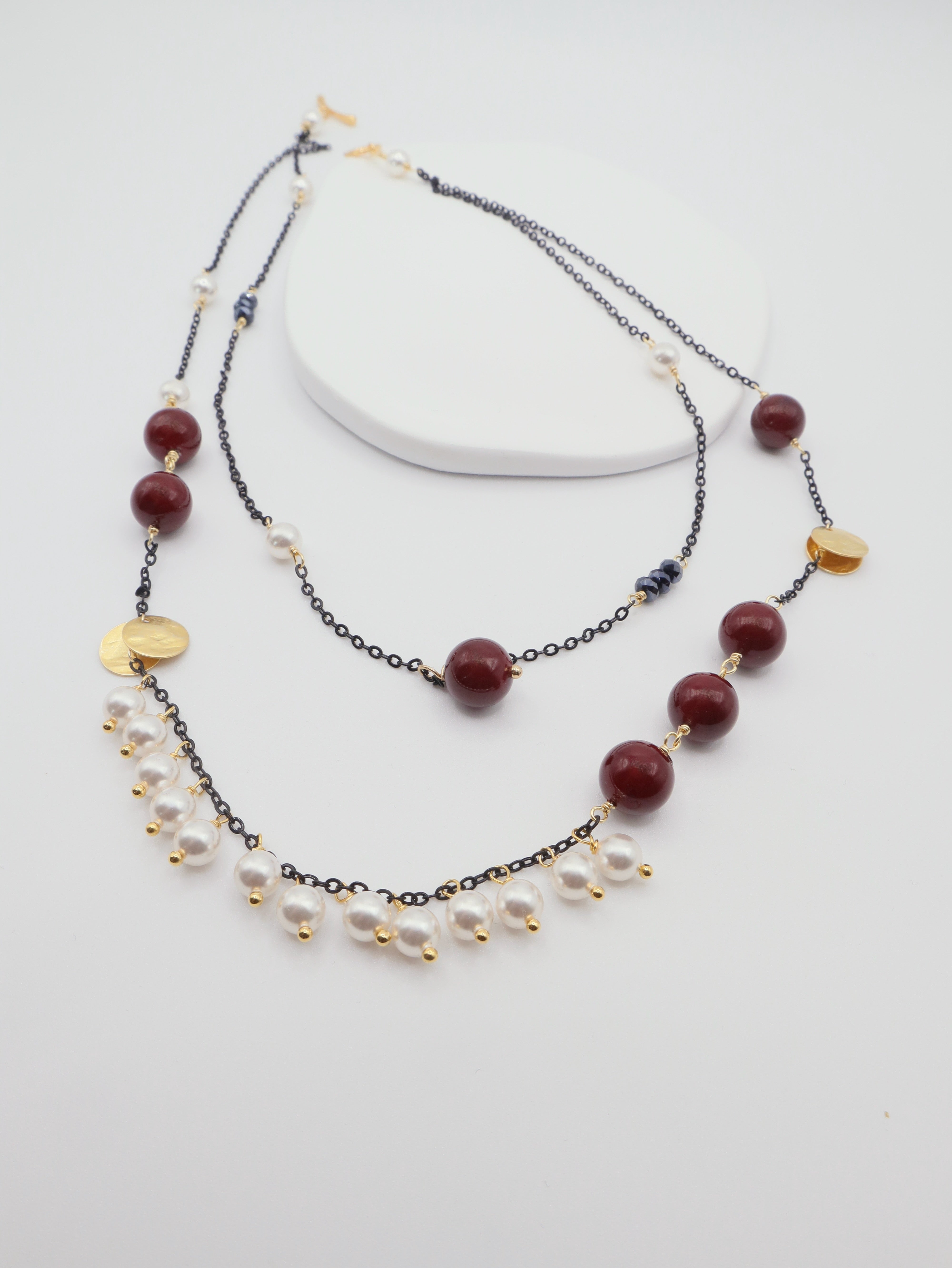 Two layered necklaces with burgundy shell pearls, black chain and gold accents on a white background